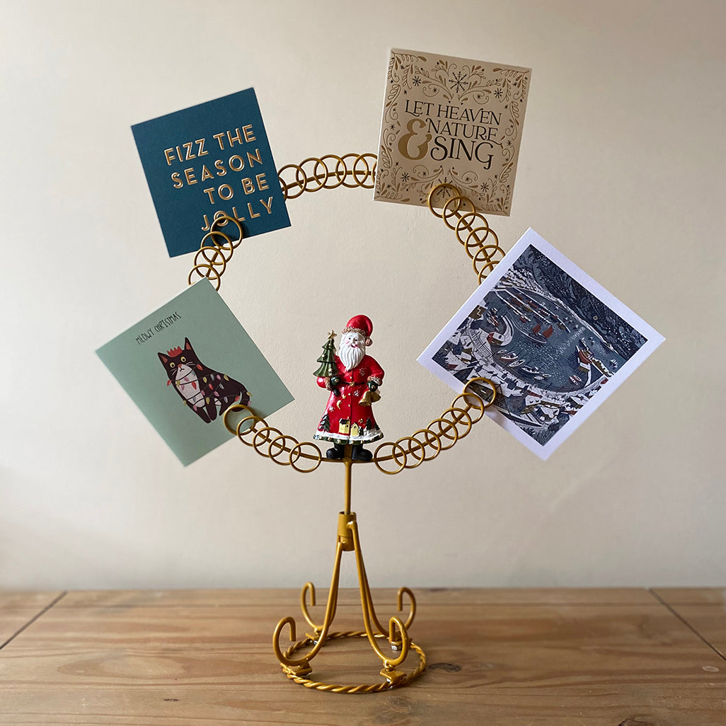 A gold-colored metal card holder in the shape of a circle with a hand-painted Santa Claus in the center, holding Christmas cards.