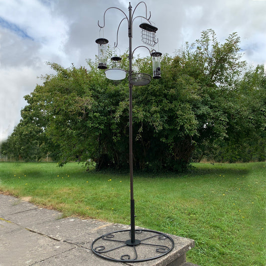 A metal bird feeding station with four feeders, a round metal patio stand, and a pole standing on grass. The feeders are bronze finished and the pole is black.