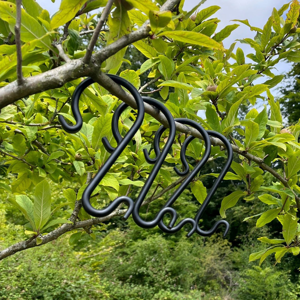 A set of black steel S hooks hanging from a tree branch surrounded by green leaves.