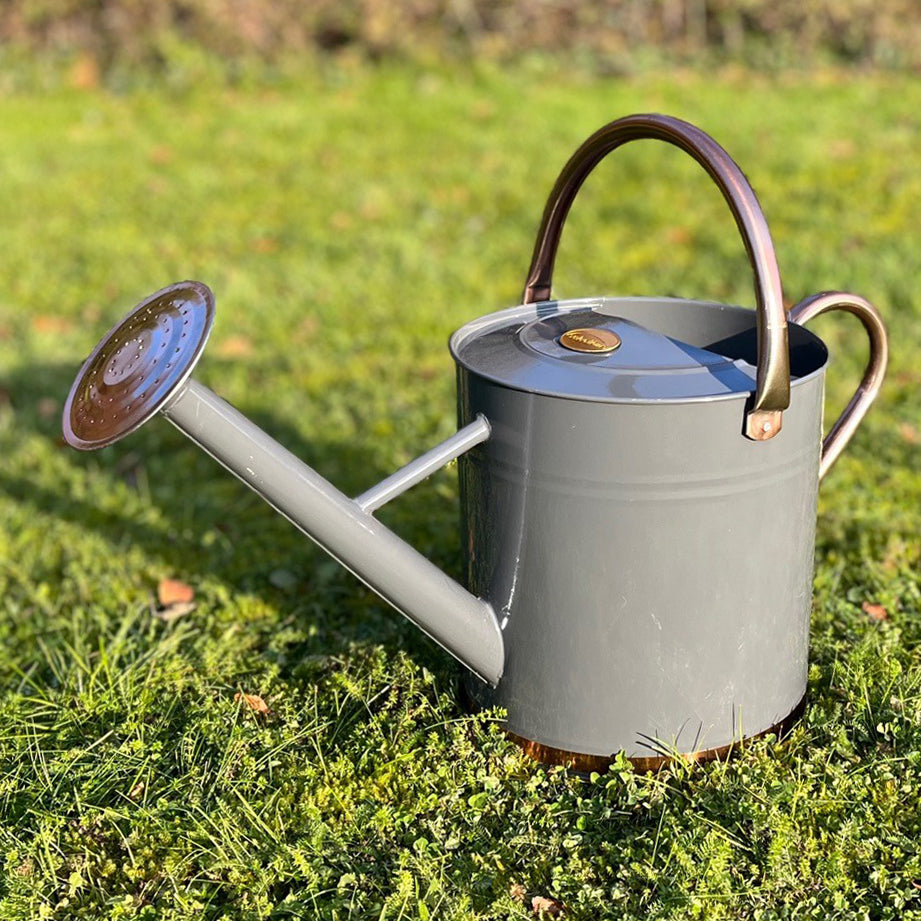 A French grey watering can with copper-colored handles and a rose detail, sitting on grass.