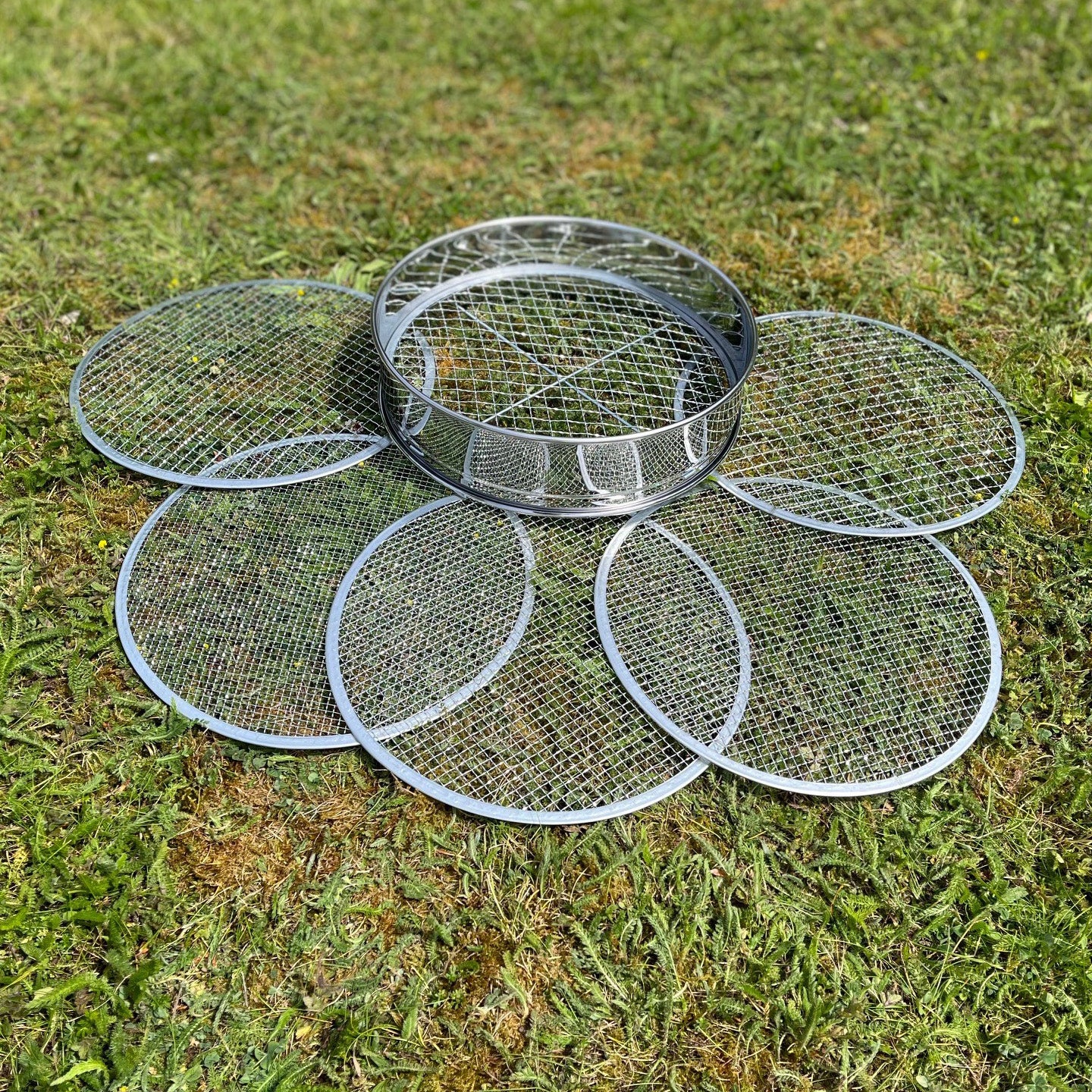 A stainless steel soil sieve with five interchangeable filters laid out on grass.