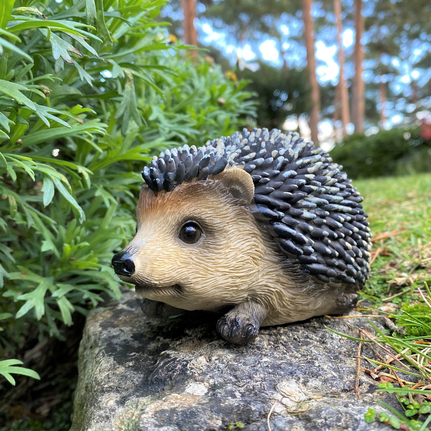 A resin garden ornament crafted into the shape of a baby hedgehog with detailed prickles and eyes, placed on a rock amidst green foliage.