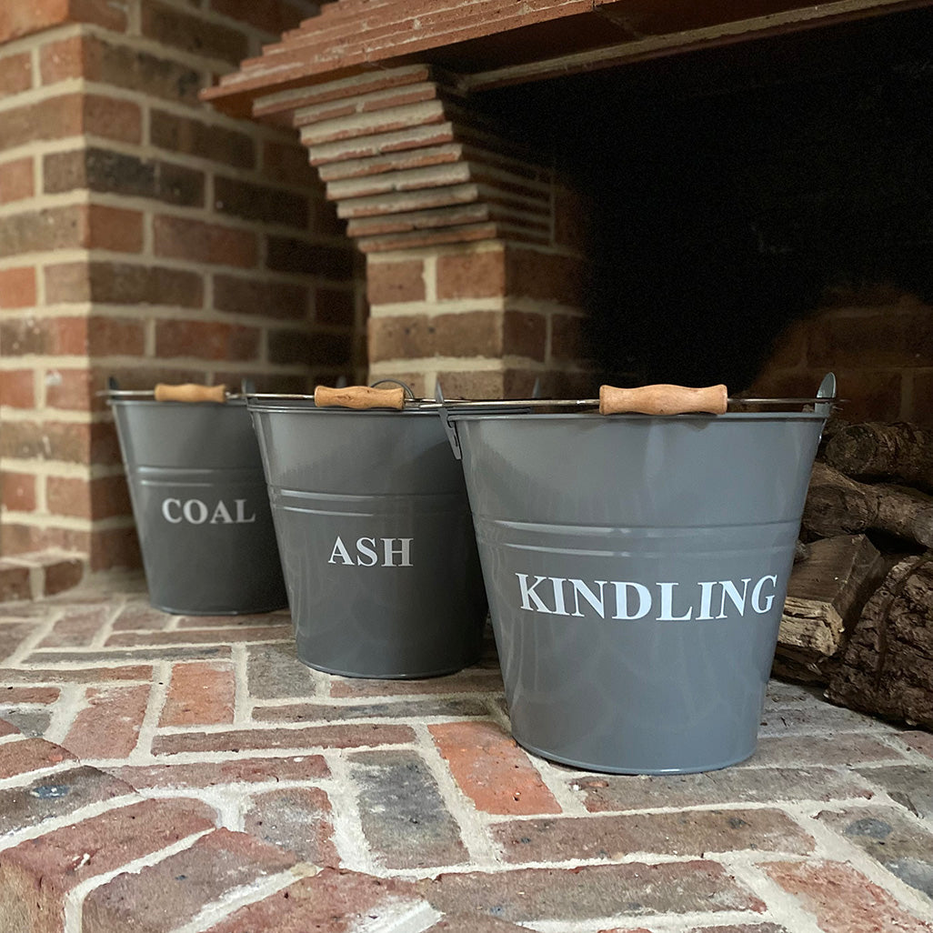 Three gray metal buckets labeled 'ASH', 'COAL', and '_KINDLING' with wooden grips on the handles, placed on a brick hearth.