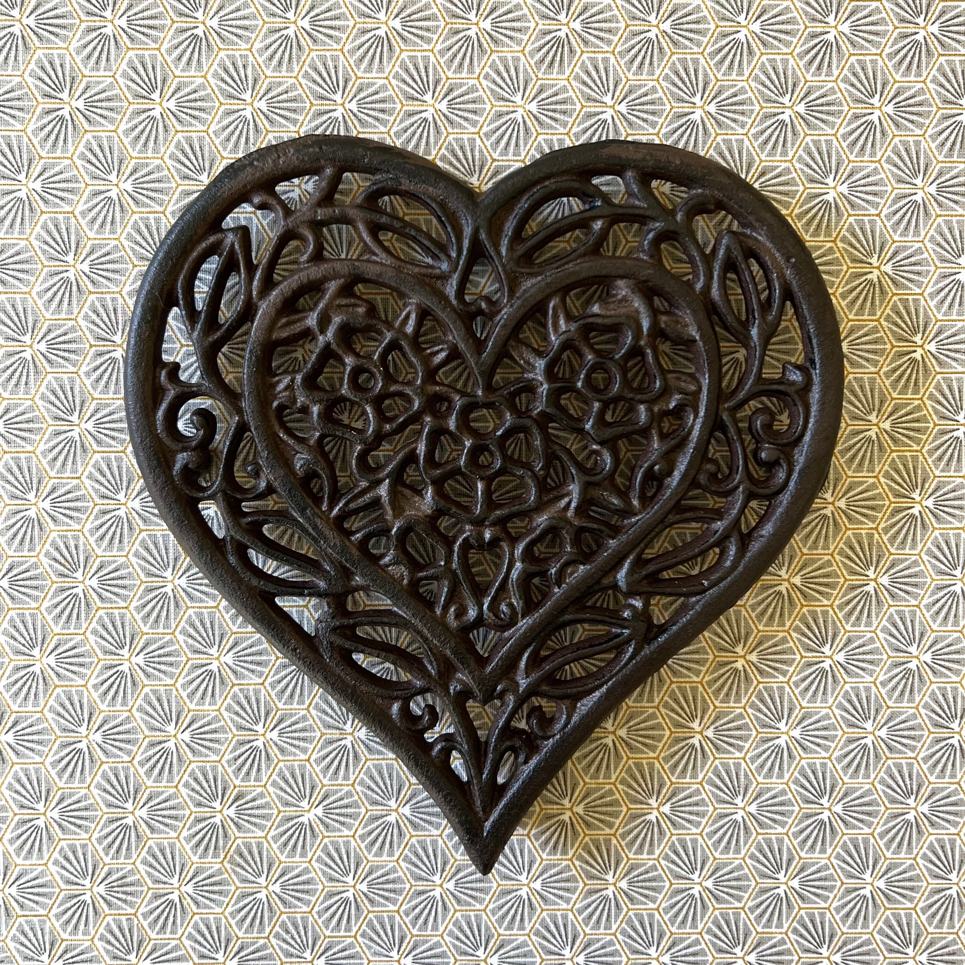 A heart-shaped black cast iron trivet with a floral internal pattern, displayed on a table with a floral design.