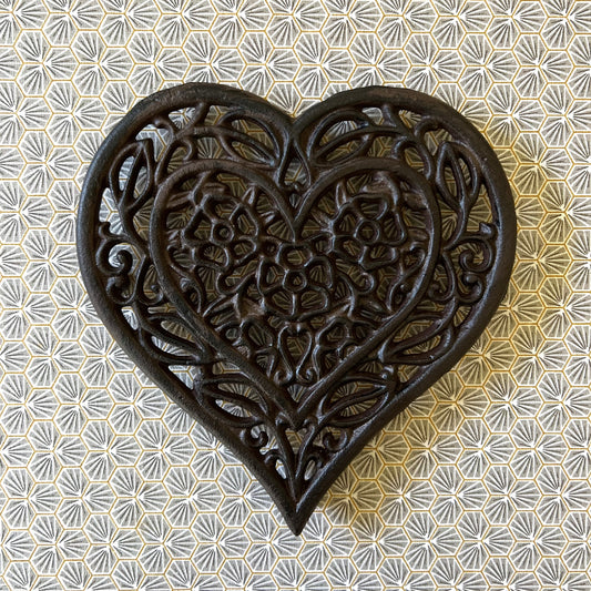 A heart-shaped black cast iron trivet with a floral internal pattern, displayed on a table with a floral design.