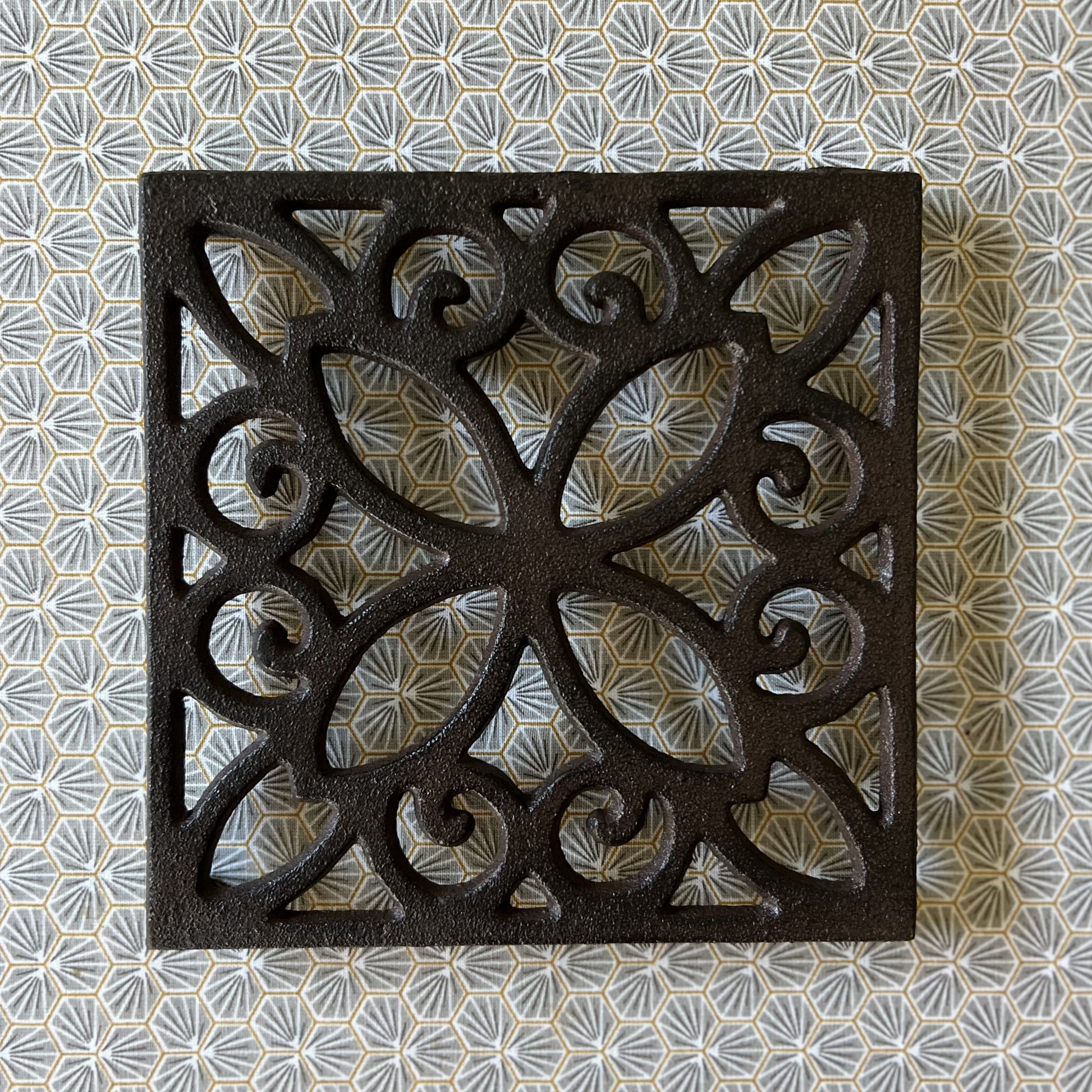 A square cast iron trivet with a vintage floral internal pattern, displayed on a patterned background.