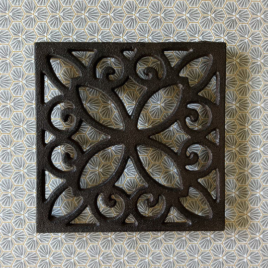 A square cast iron trivet with a vintage floral internal pattern, displayed on a patterned background.