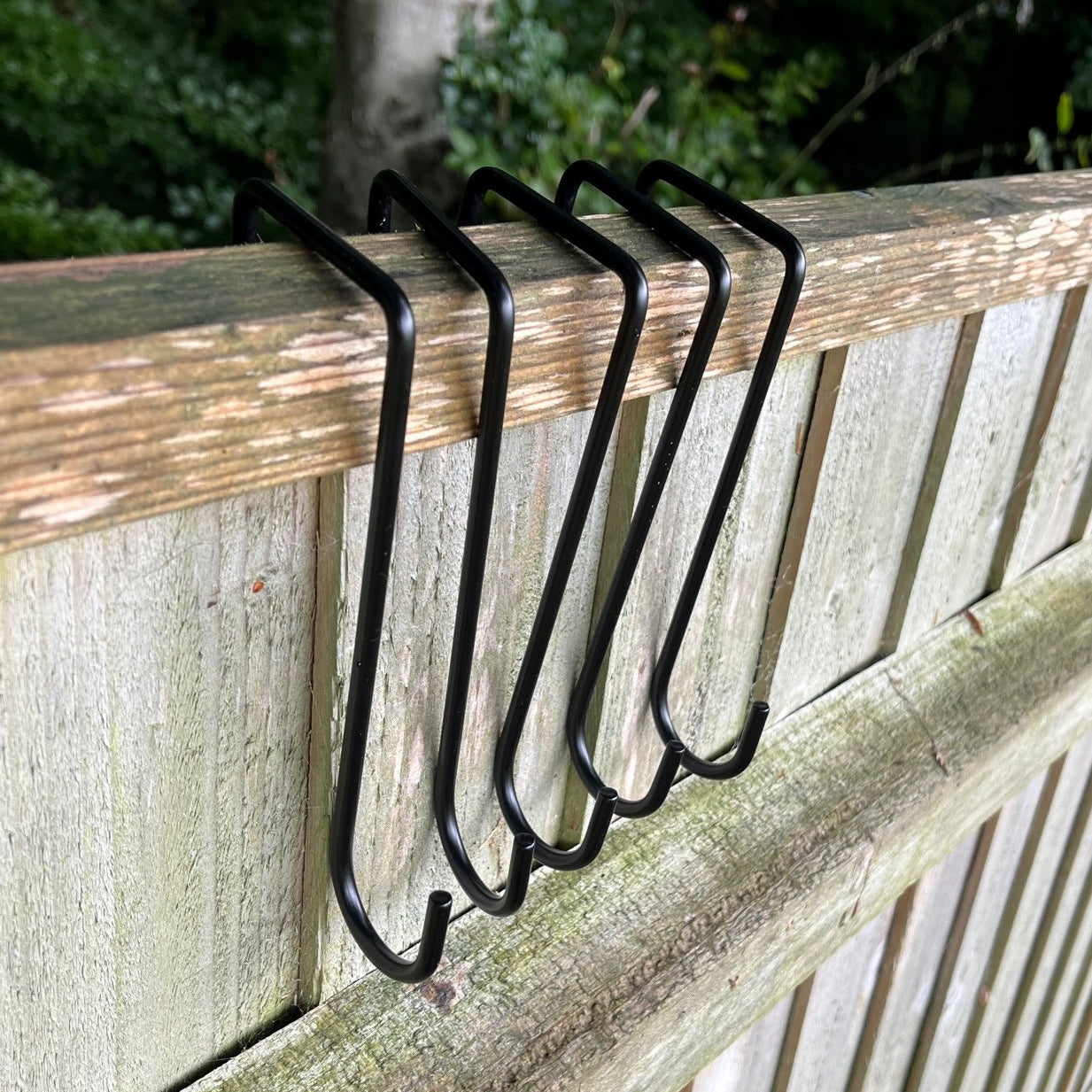 A set of black painted steel hooks slots on a wooden fence panel, designed for hanging planters or decorations.