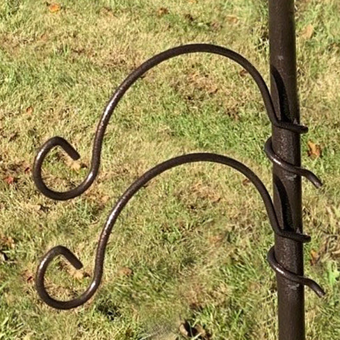 A black metal hanging bracket hook with spiral fixings attached to a pole, set against a grassy background.