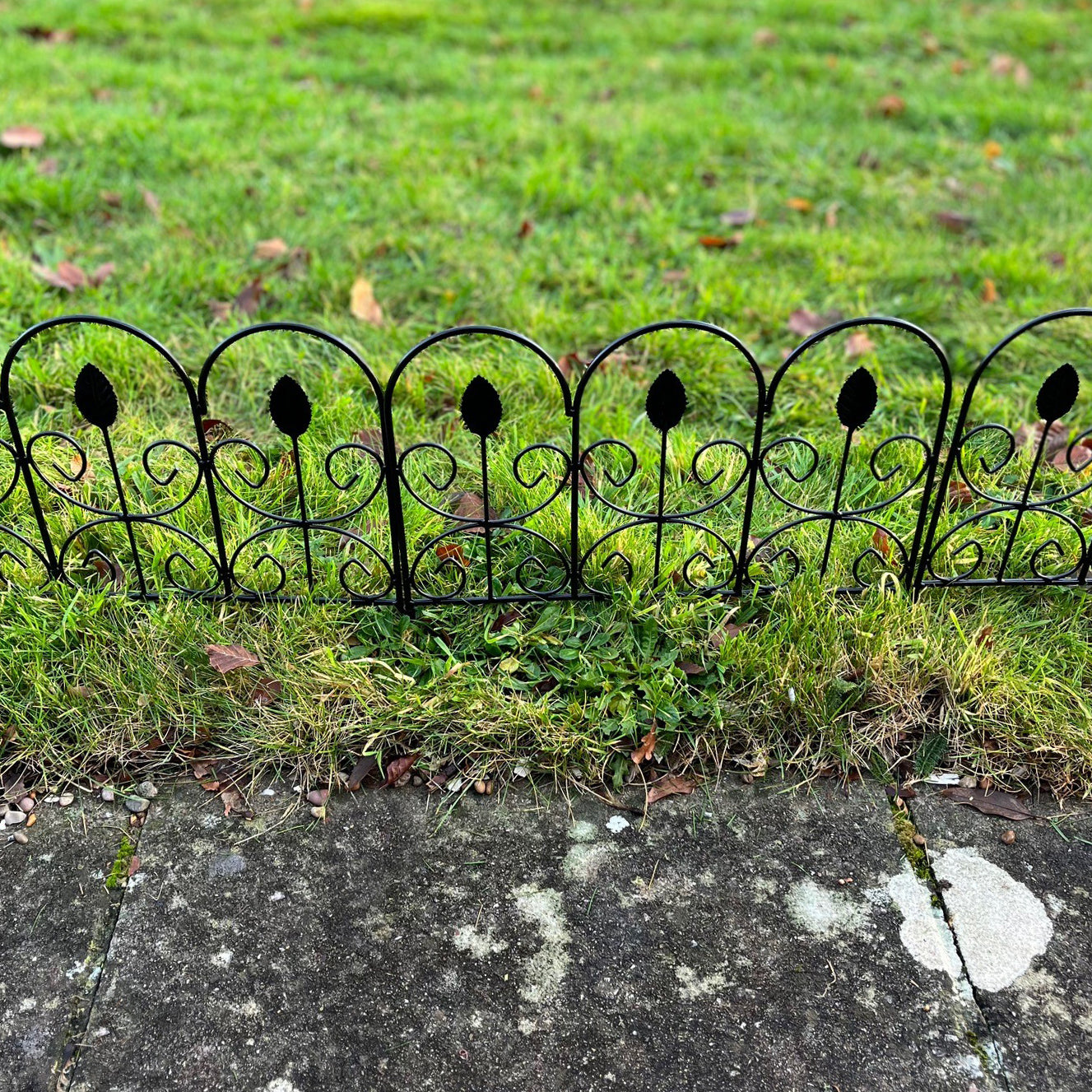 A black metal garden lawn edging panel with a leaf design, placed on a grass border next to a concrete path.