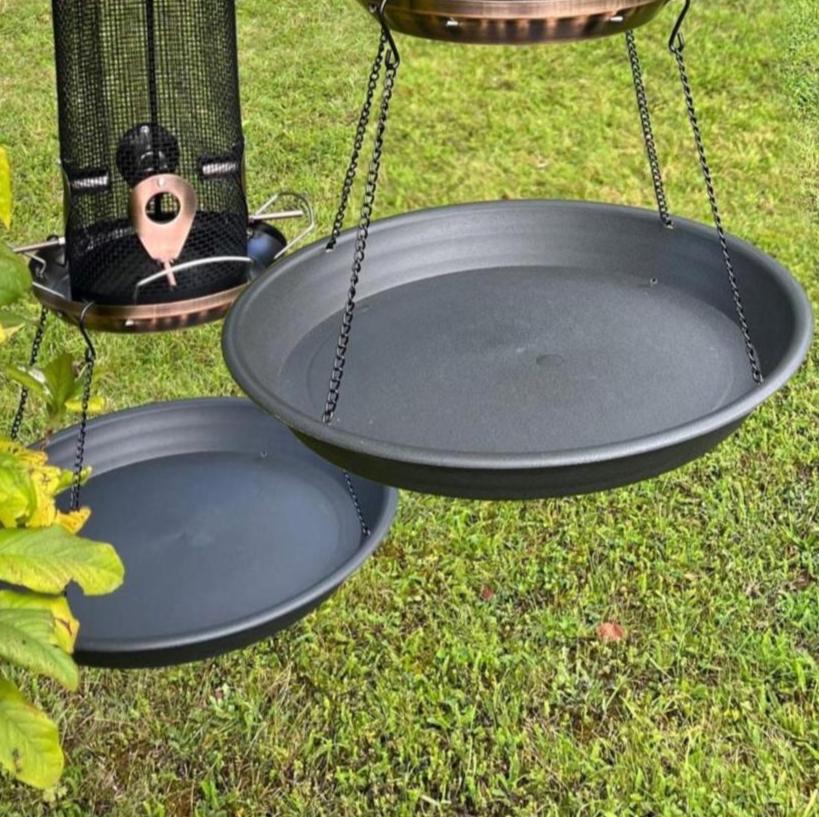 Two black plastic trays attached to bird feeders by chains, designed to catch dropped seeds, hanging over grass.