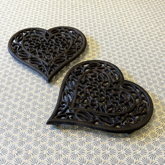 Two black heart-shaped cast iron trivets with floral internal patterns, placed on a table with a similar floral pattern.