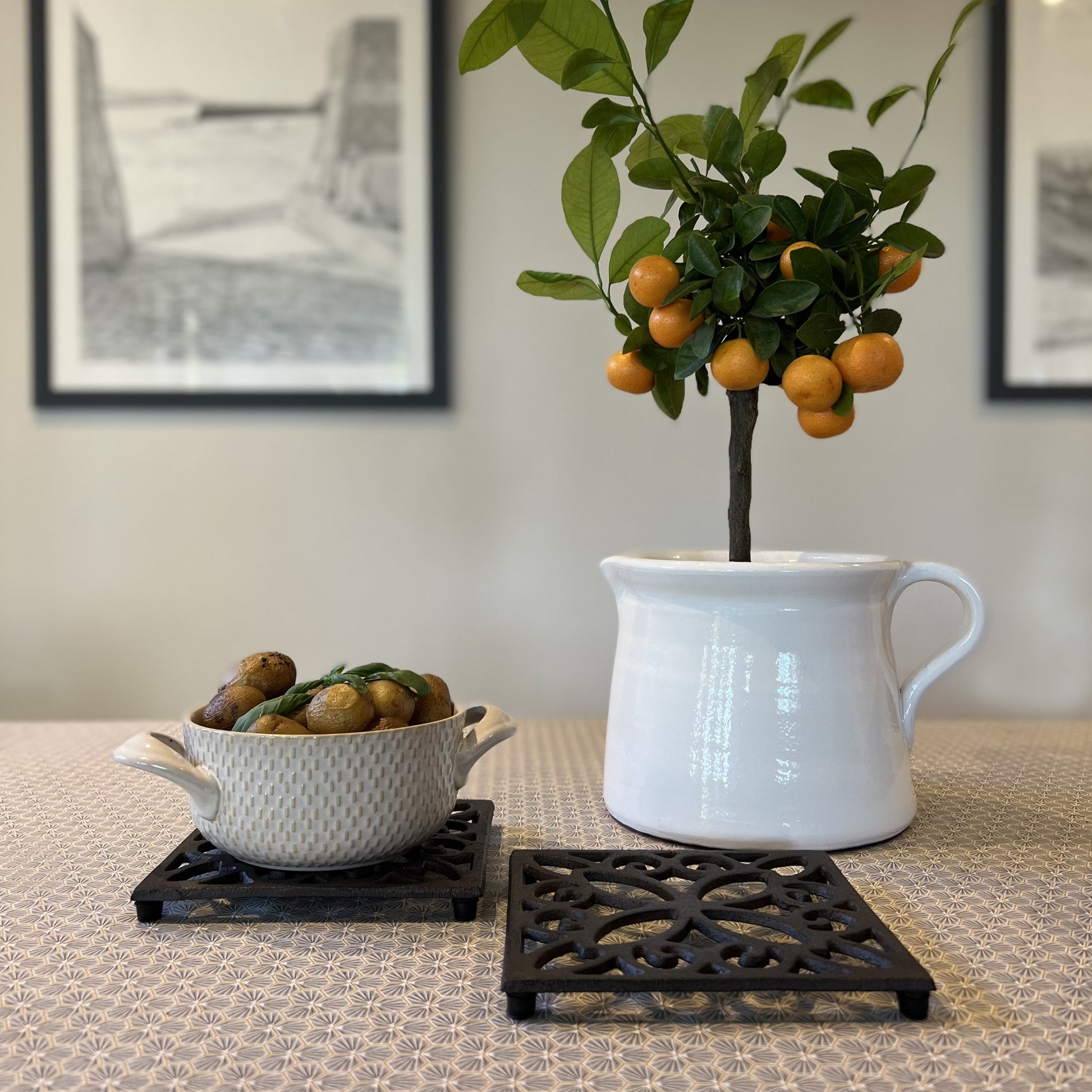 Two square black cast iron trivets with a vintage pattern, placed on a table with a white vase on top of one of them and a bowl on the other, set against a backdrop of a framed black and white photograph of a tree.