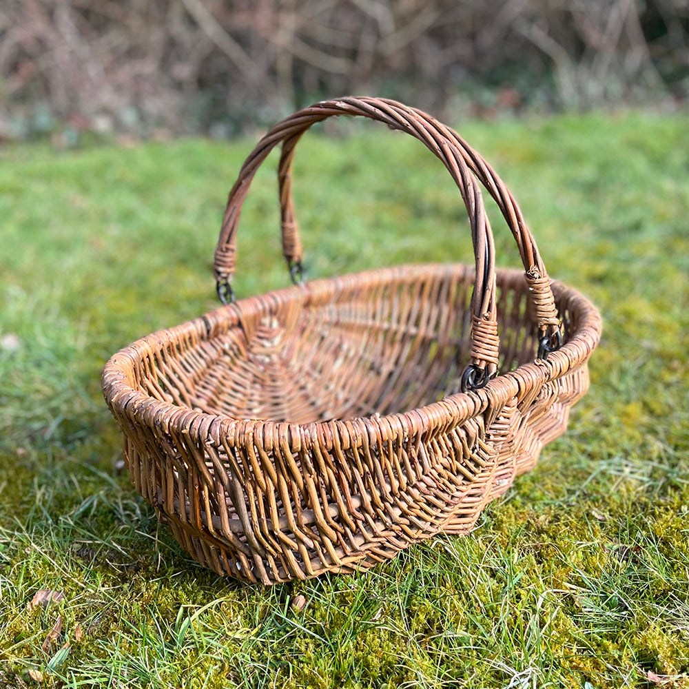 A rustic wicker garden trug basket with folding handles resting on grass.
