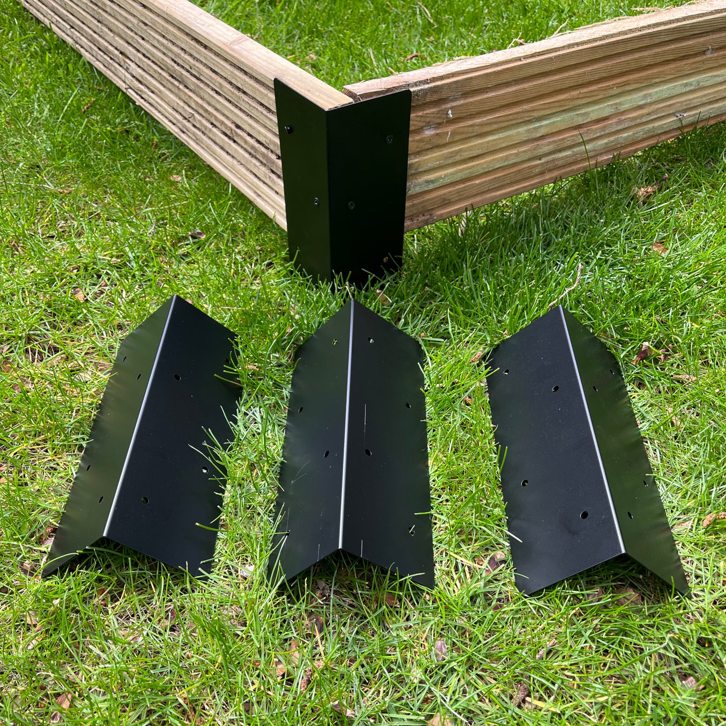 Black steel raised bed corner brackets displayed on grass alongside a partial view of a wooden raised garden bed.