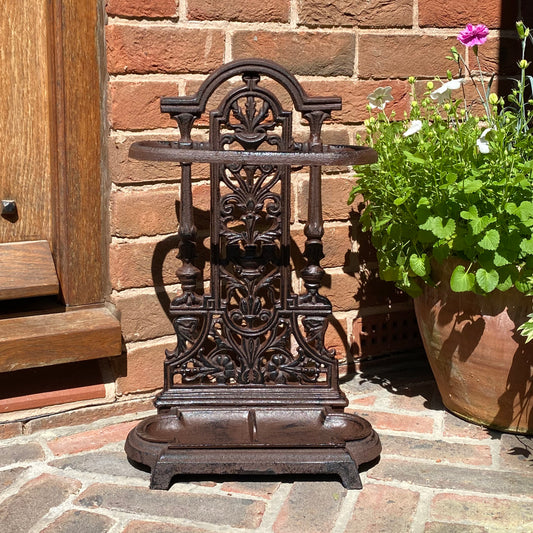 A vintage style brown cast iron umbrella stand placed on a brick floor next to a potted plant.