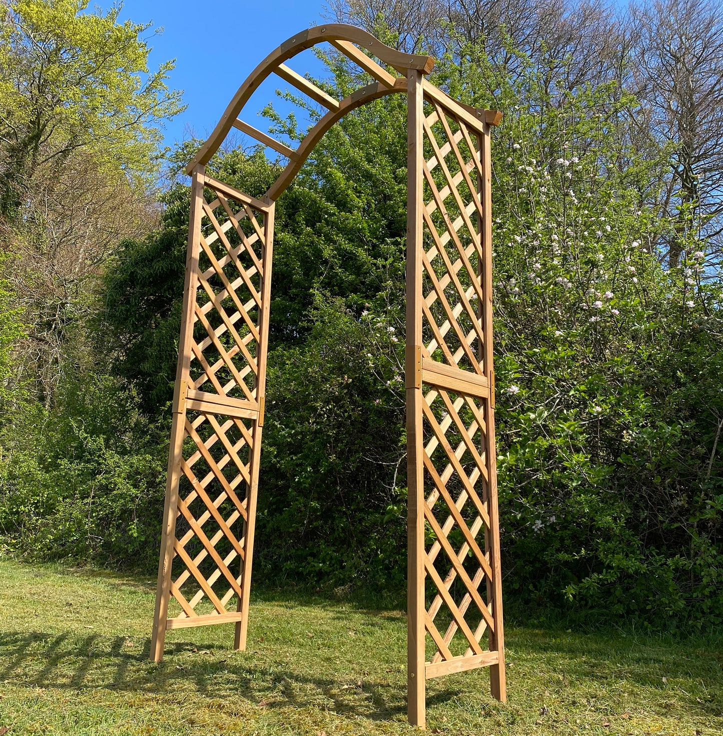 A wooden garden arch with a curved top and trellis sides, standing on grass with trees in the background.