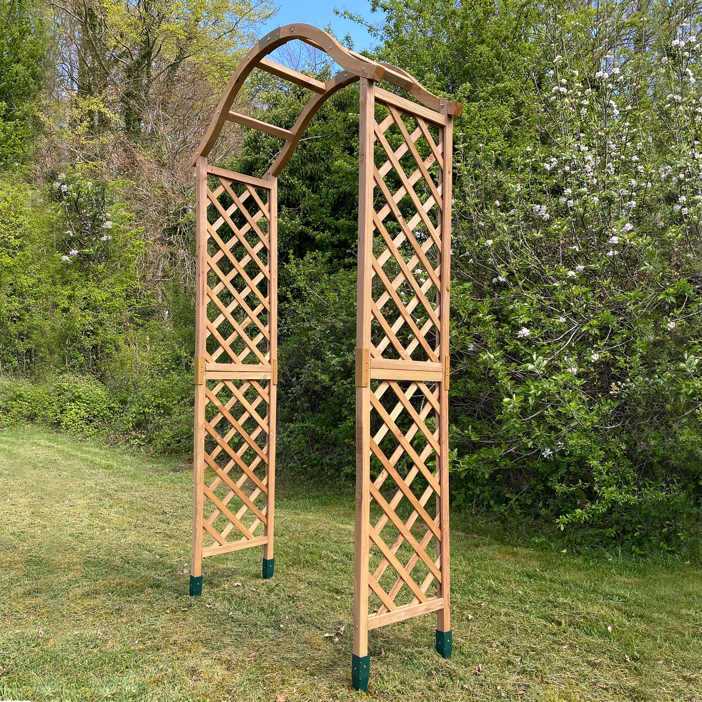 A wooden garden arch with trellis sides and a smoothly curved top, supported by four sturdy feet, installed on a grassy area with trees in the background.