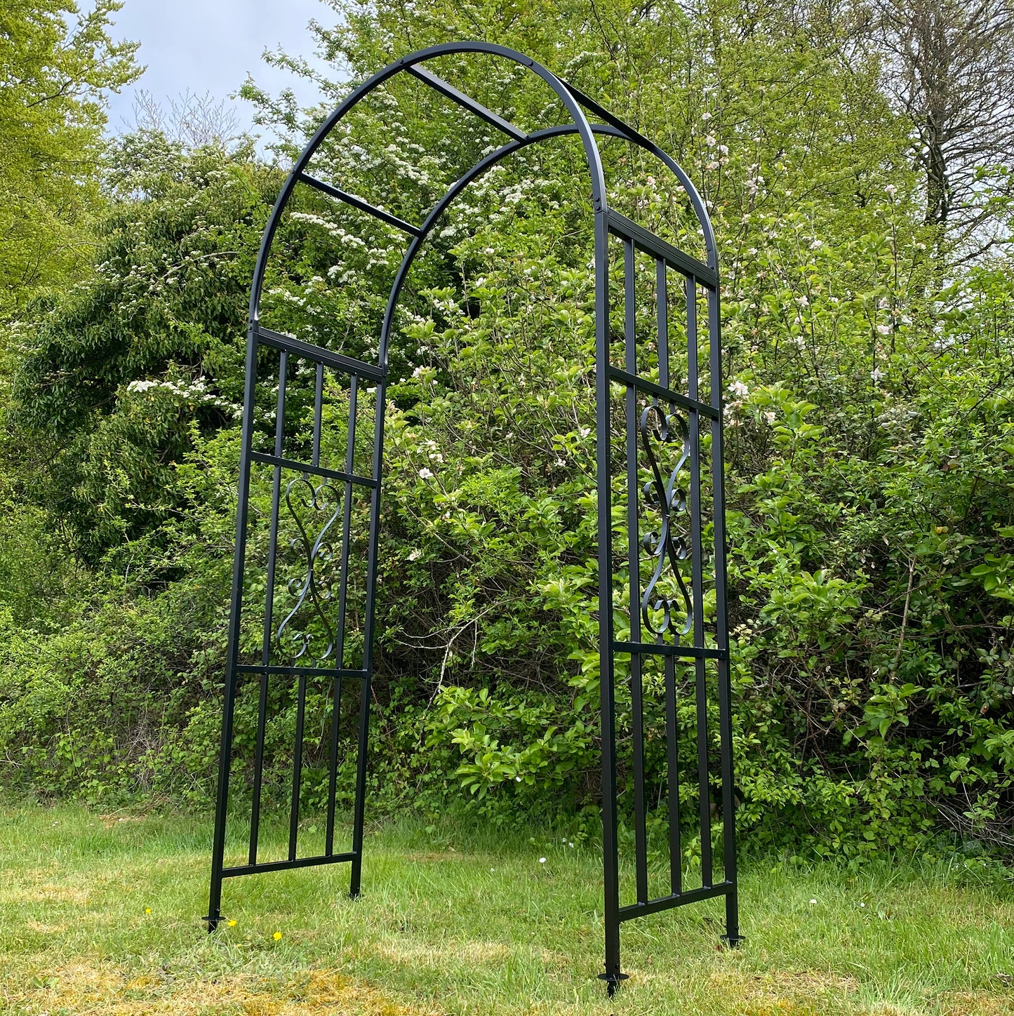 A black metal garden arch installed in a garden setting with greenery in the background.