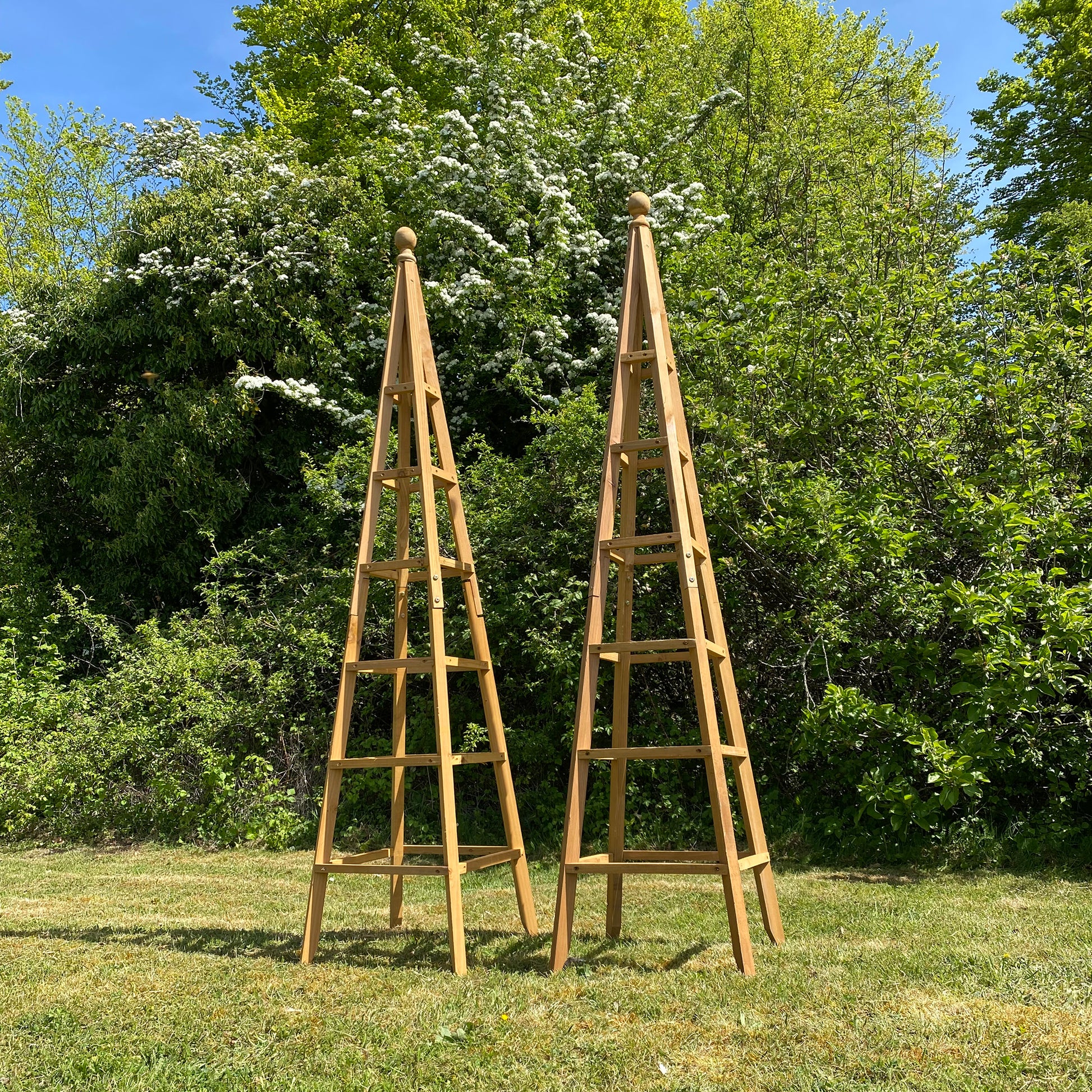 Two wooden garden obelisks standing on grass with trees in the background, classic simple design with an attractive top finial.