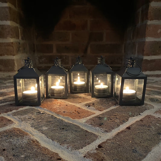 A set of five metal and glass candle lanterns with tealight candles inside, placed on a brick surface.