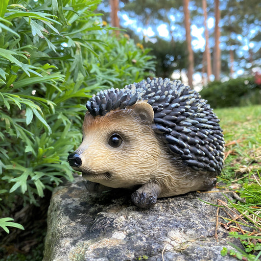 A resin garden ornament crafted into the shape of a baby hedgehog with detailed prickles and eyes, placed on a rock amidst green foliage.